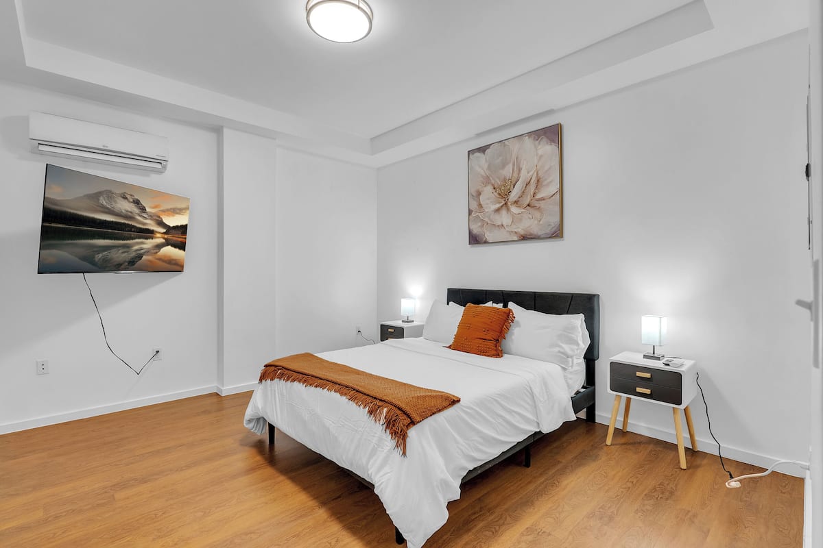 A bedroom with a bed, nightstands, a wall-mounted air conditioner, and artwork.
