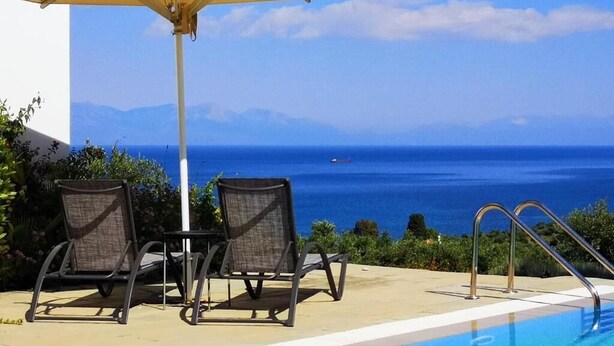 A poolside area with two chairs and an umbrella, overlooking a clear blue sea and distant mountains.