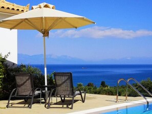 Zona junto a la piscina con dos sillas y una sombrilla, con vistas al mar azul y montañas al lejano.