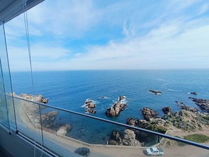 Vista costeira de um ponto elevado, com afloramentos rochosos no mar, céu claro e uma estrada com veículos.