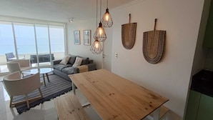 A modern living room with a wooden dining table, grey sofa, and a large window offering a view. - Apartment in Concon, in front of the sea. Wonderful view. 5 people, very complete. (Concón)
