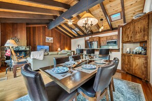 A rustic interior with a dining table set for six, wooden beams on the ceiling, a fireplace, and a kitchen area. - Chinquapin Escape | Pool + Sauna + 2 Beaches (Carnelian Bay)
