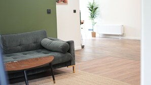 Een kamer met een grijze bank, een houten bijzettafeltje, een groene accentmuur, een radiator en een kamerplant.
