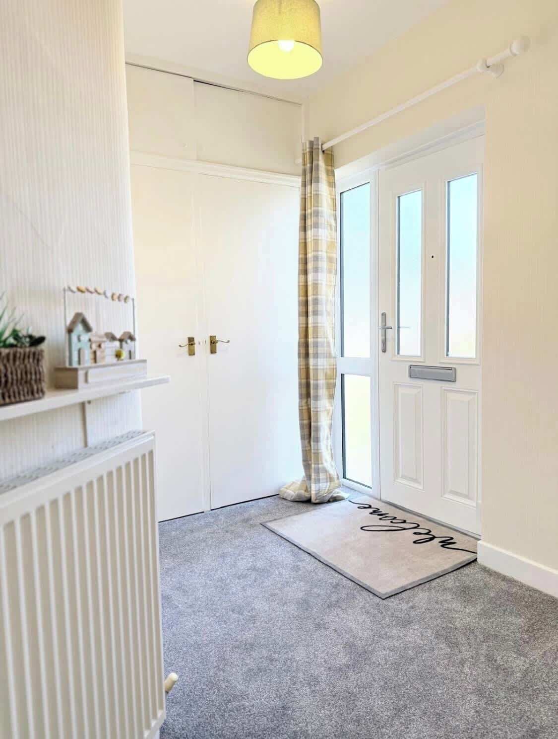 A hallway with a white door, a radiator, a small shelf with decorative items, and a doormat with a name.