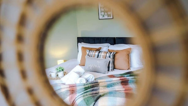 Ein Bett mit einer Karo-Decke, mehreren Kissen, einem Nachttisch mit Tasse und Pflanze sowie einem gerahmten Bild an der Wand.