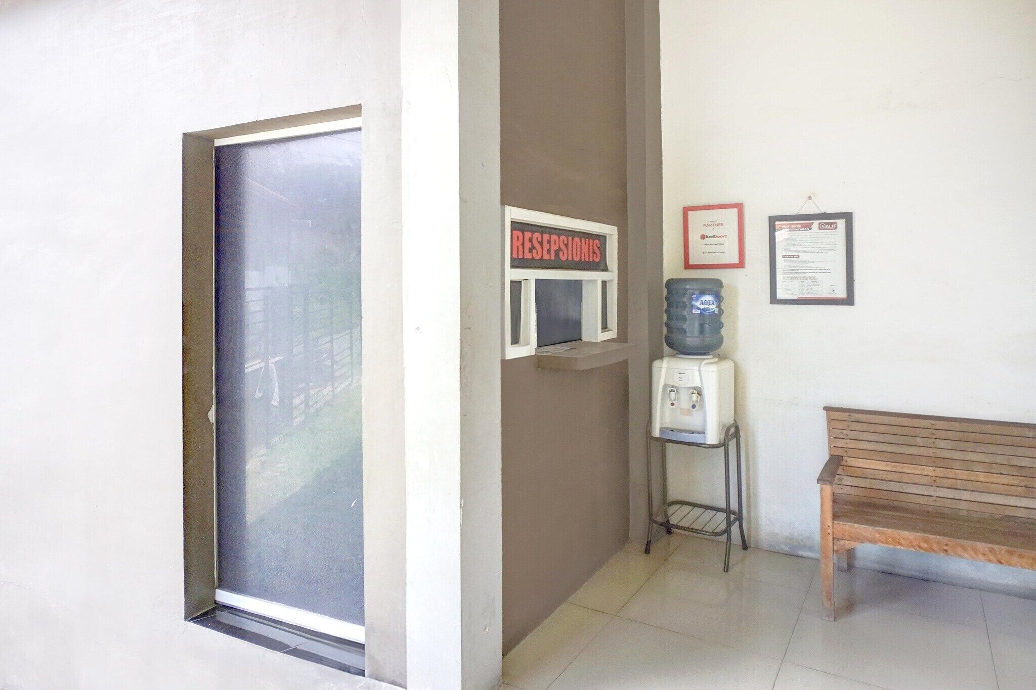 A reception area with a sign reading 'RECEPTIONIS,' a water dispenser, a bench, and a framed document on the wall.