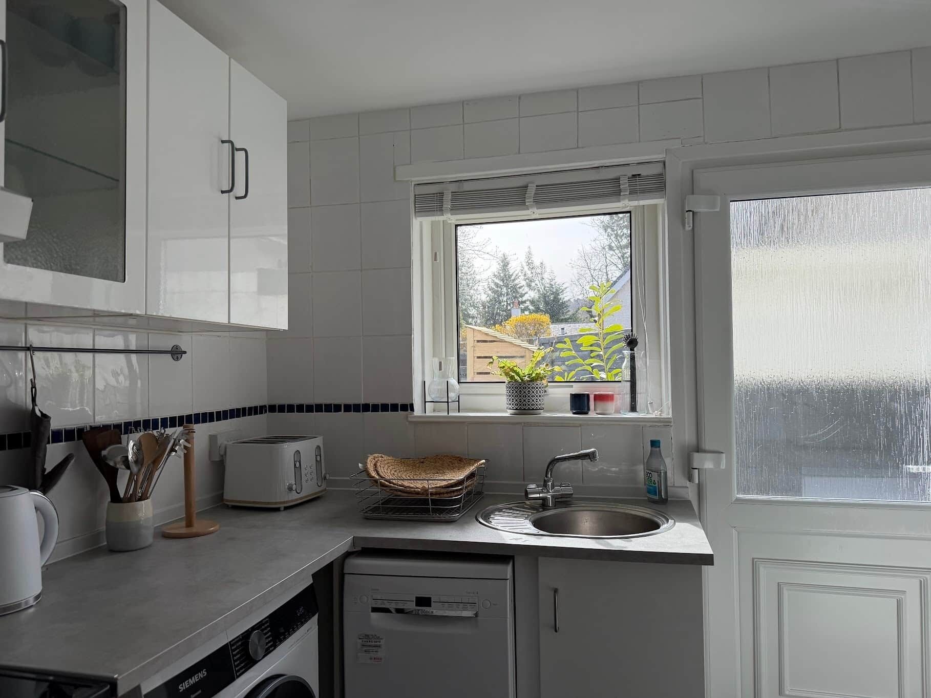 A kitchen with white cabinets, a window with a view of trees, a sink, and a dishwasher.
