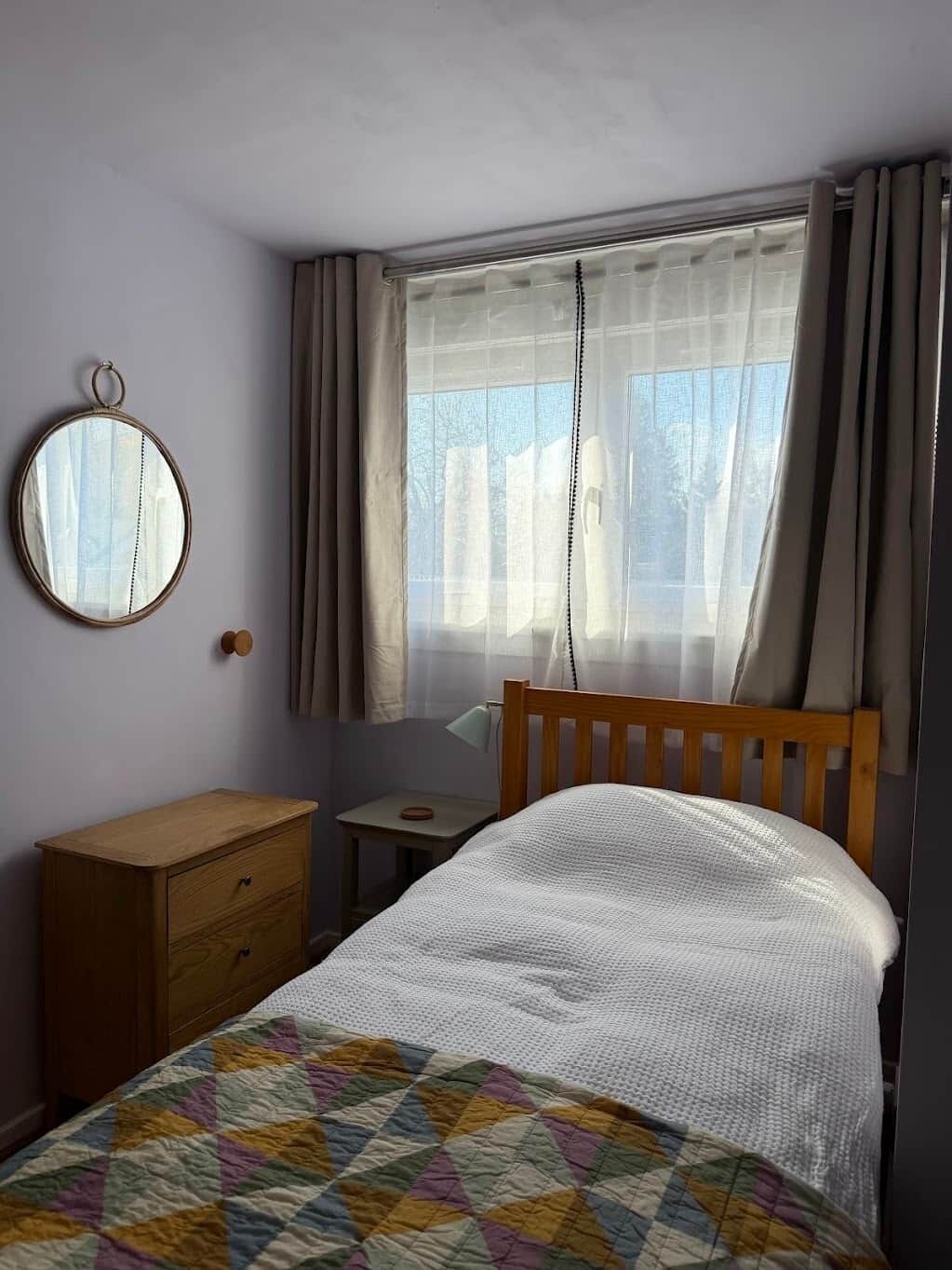 A bedroom with a bed, a wooden dresser, a small table, a mirror, and a window with curtains.