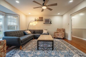 A living room with a sectional sofa, a coffee table, patterned rug, ceiling fan, and wall-mounted lamps.