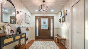 Un pasillo con suelo de madera, alfombra de cuadros, una consola decorada con objetos, un espejo, apliques en la pared y una puerta con paneles de vidrio esmerilado.