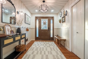 A hallway with a wooden floor, a checkered rug, a console table with decorative items, a mirror, wall sconces, and a door with frosted glass panels.