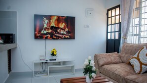 Ein Wohnzimmer mit Flachbildfernseher, einer Couch, einem Couchtisch und einem kleinen weißen Regal mit einer Vase voller Blumen.
