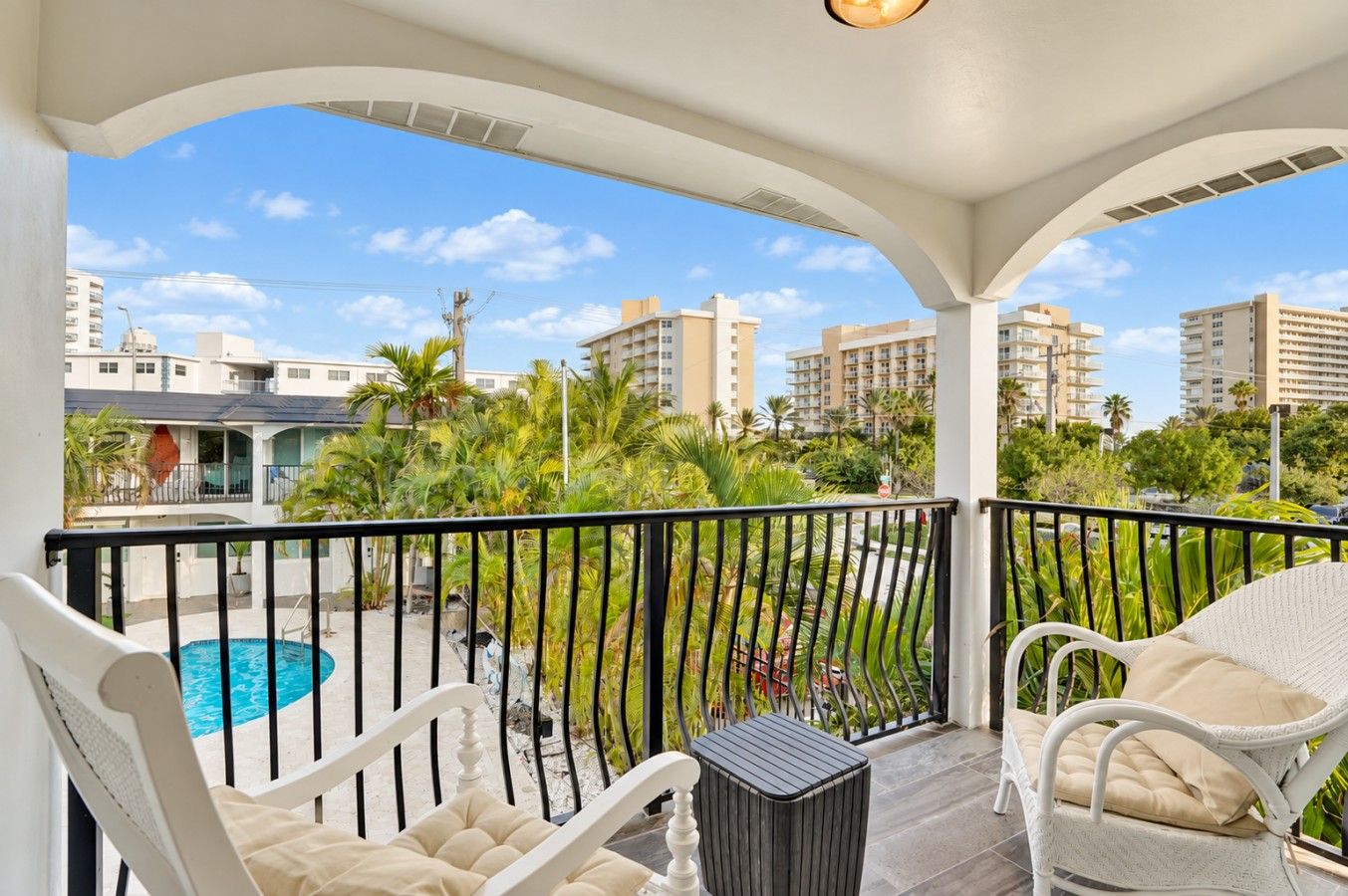 A balcony with a pool, white wicker furniture, and a view of surrounding buildings and greenery.