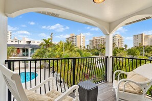 A balcony with a pool, white wicker furniture, and a view of surrounding buildings and greenery. - 3BR Beach Retreat | Heated Pool + Parking (Pompano Beach)