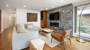 Un soggiorno moderno con un divano grigio, un tavolino in legno, una poltrona in pelle, una TV a schermo piatto montata su una parete in pietra e un camino.