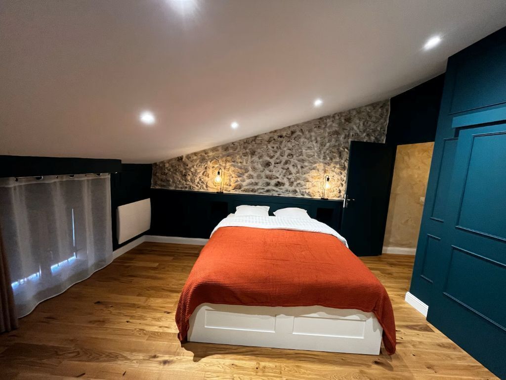 Ein Schlafzimmer mit einer Steinwand, einem Bett mit orangefarbener Decke, Holzboden und einer türkisblauen Tür.