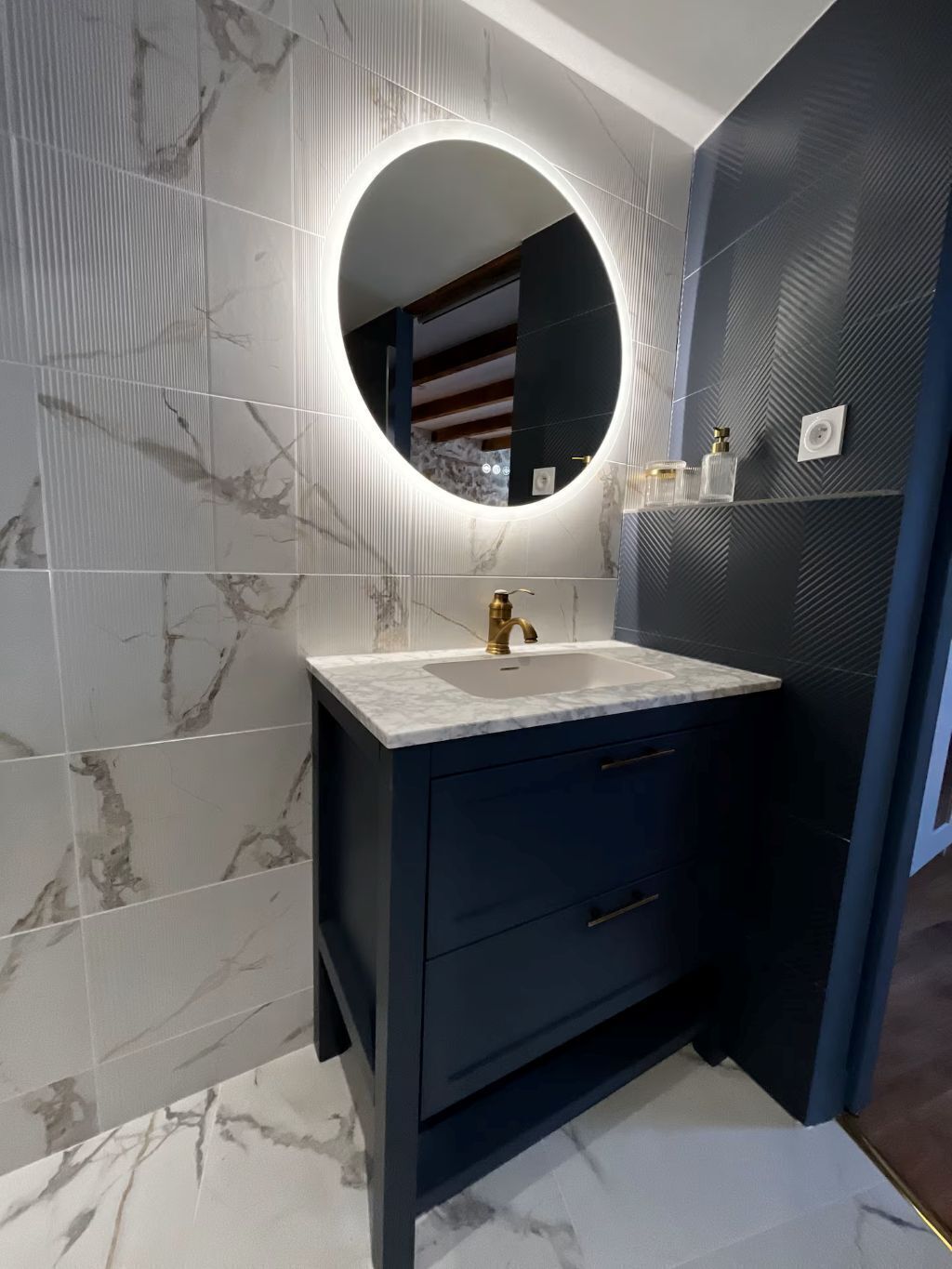 A bathroom with a marble-tiled wall, a dark vanity with a gold faucet, and a round mirror with a light.