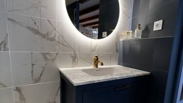A bathroom with a marble-tiled wall, a dark vanity with a gold faucet, and a round mirror with a light.