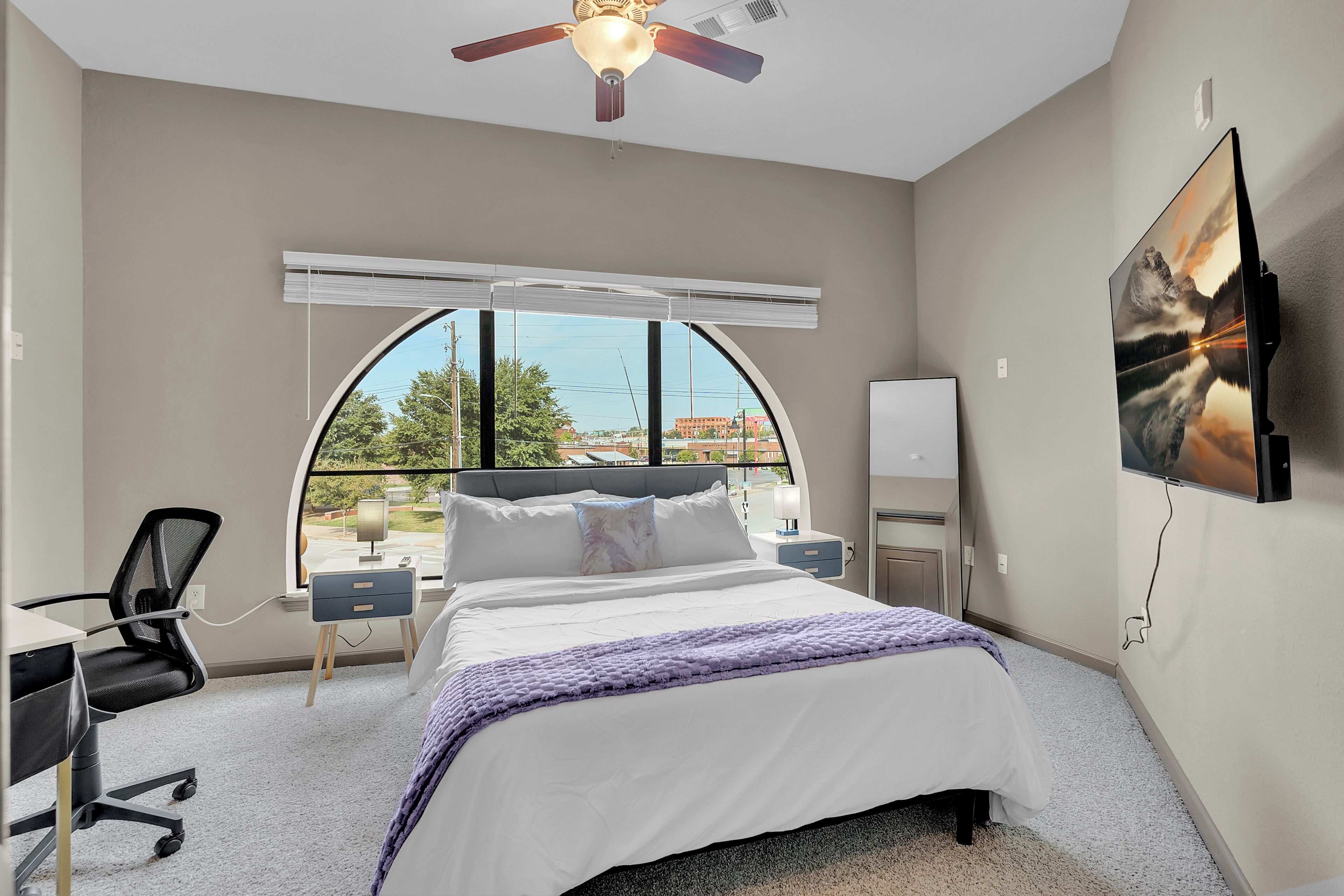 A bedroom with a bed, desk, chair, and a large window with a view.