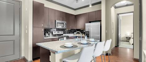 Una cocina moderna con armarios de madera oscura, electrodomésticos de acero inoxidable, encimera de granito y una zona de bar con sillas blancas.