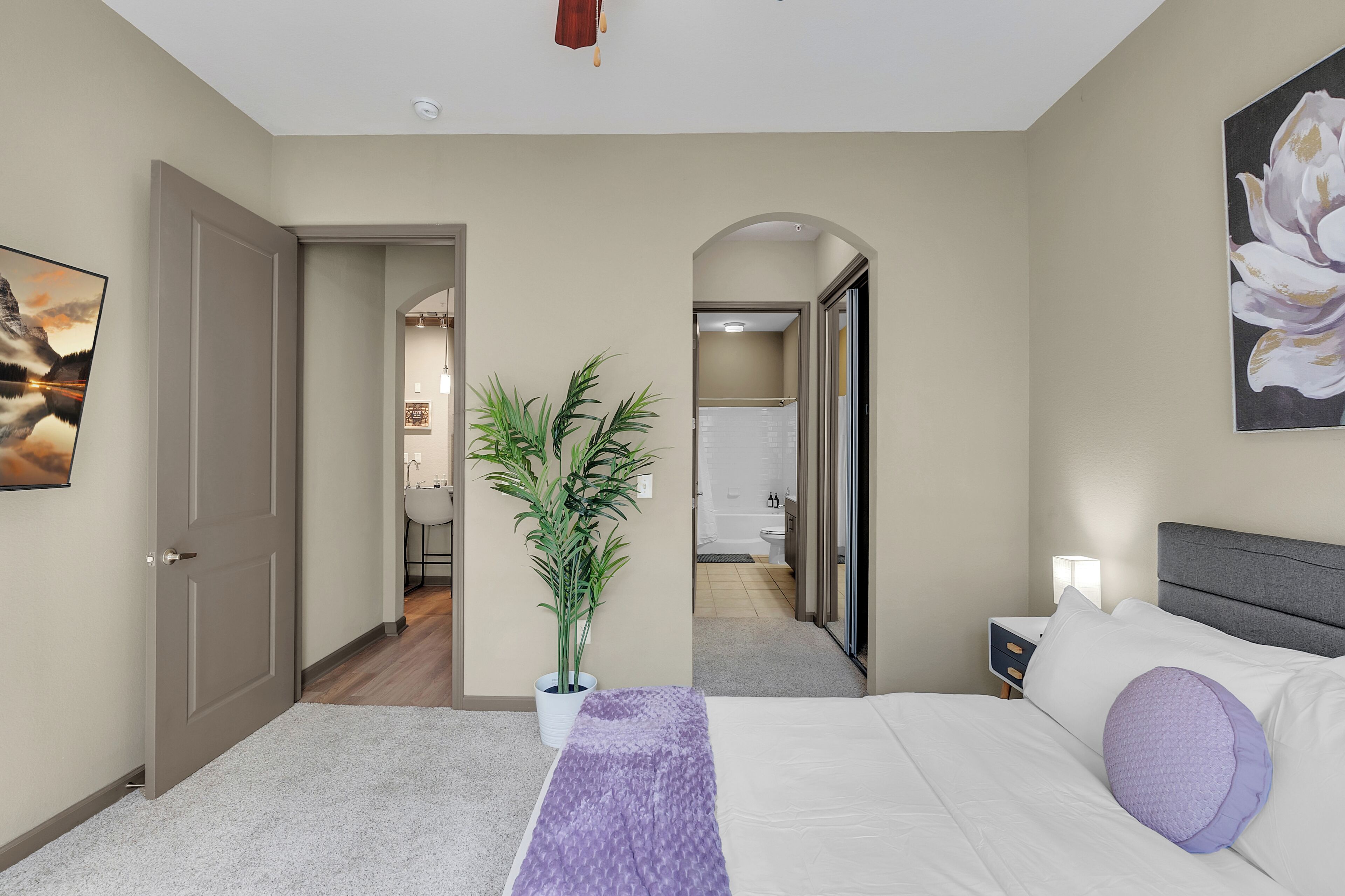 A bedroom with a bed, a nightstand, a potted plant, and a hallway leading to a bathroom.
