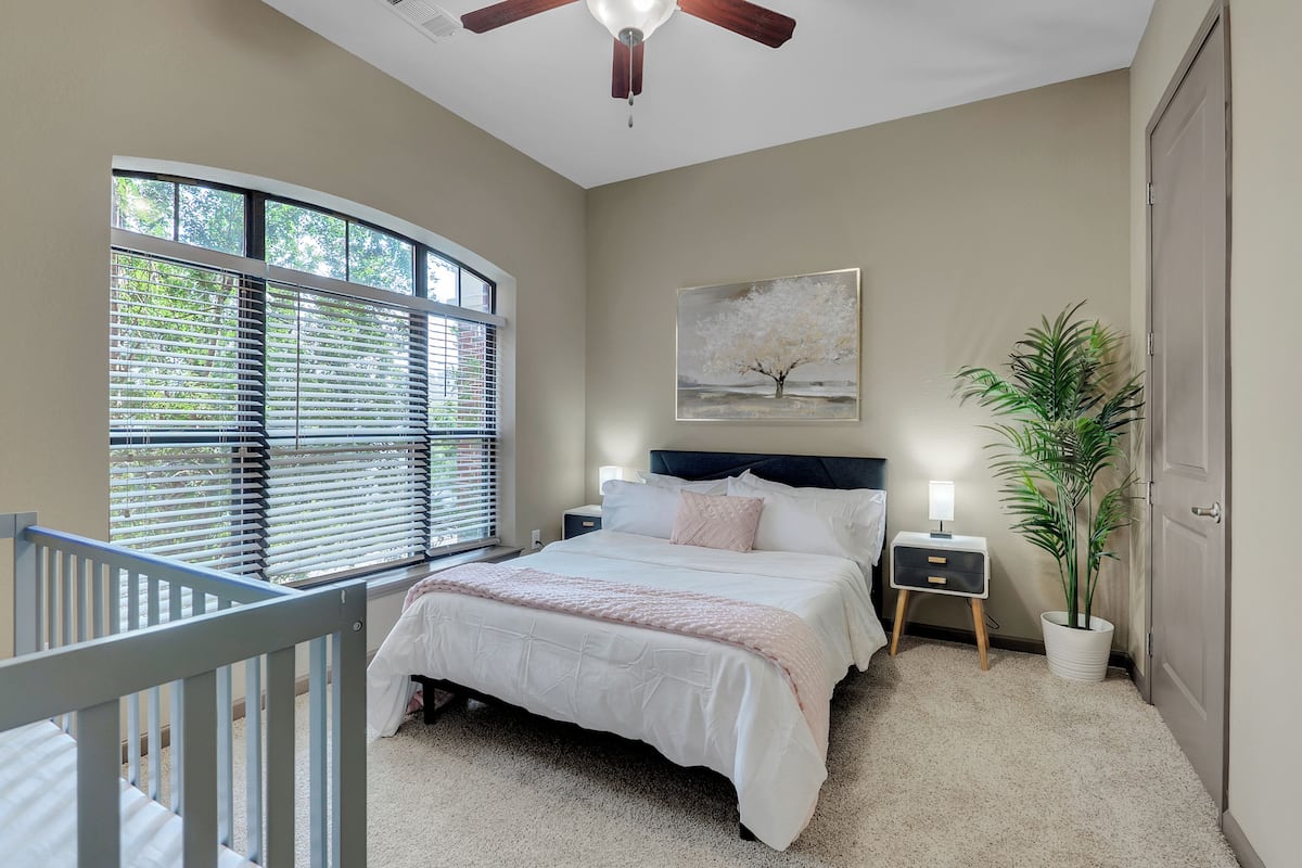 A bedroom with a bed, nightstands, a ceiling fan, a window with blinds, a painting, and a potted plant.