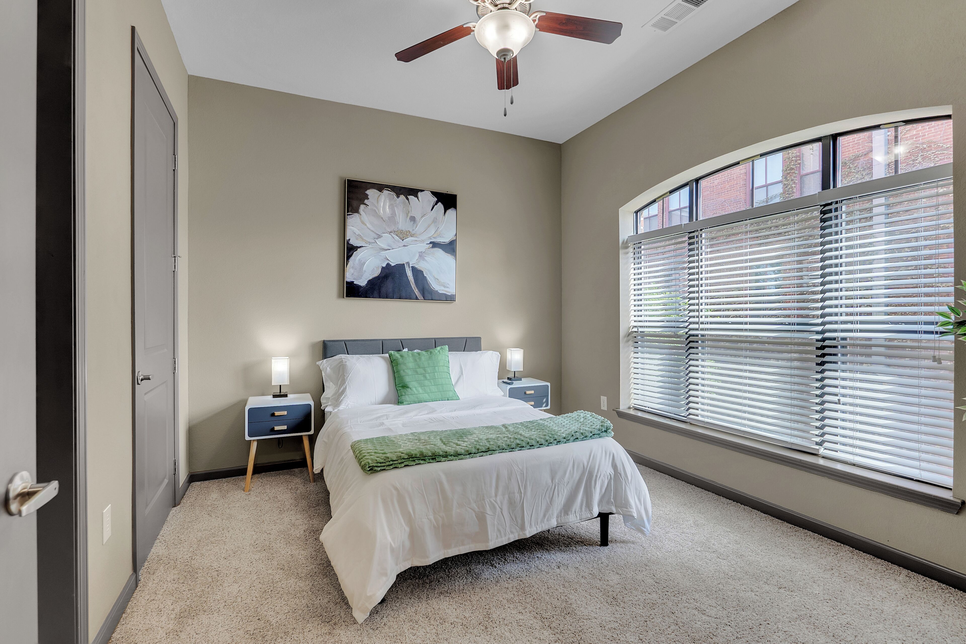 A bedroom with a bed, a ceiling fan, a nightstand, a bedside table, a window with blinds, and a wall-mounted painting.