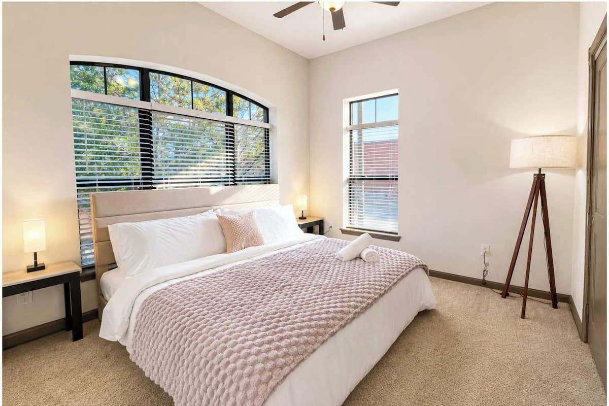 A bedroom with a bed, two bedside tables, a floor lamp, and a window with blinds.