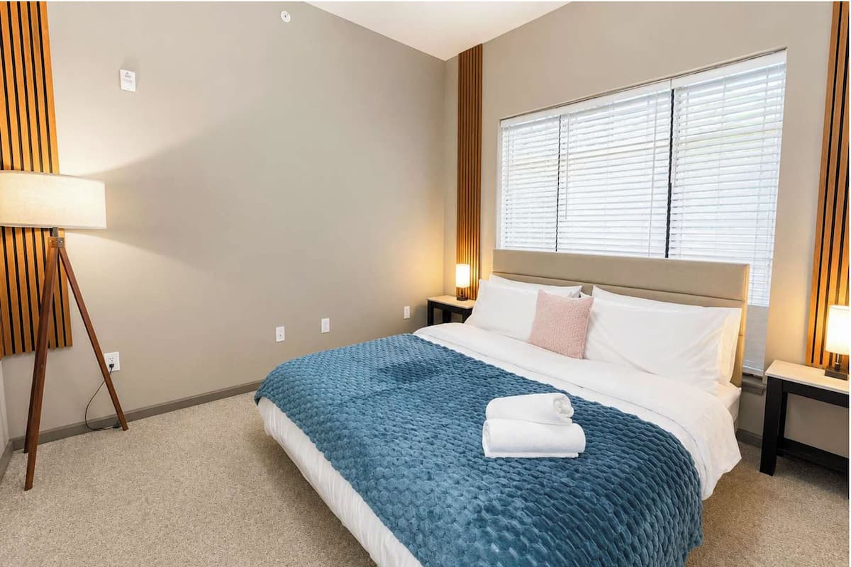 A hotel room with a bed, a lamp, a window with blinds, and a wall-mounted light fixture.