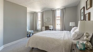 Un dormitorio con una cama grande, una mesita de noche con lámpara, una silla y una ventana con cortinas.