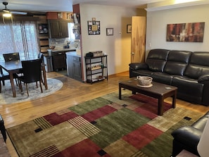 A living room with a sofa, coffee table, and a rug. There is a dining area with a table and chairs. A kitchen is visible in the background.