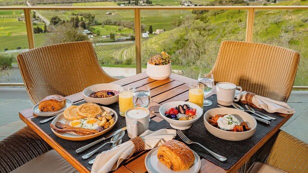 Una mesa de desayuno con diversos platos, vista a un paisaje verde y sillas de mimbre.