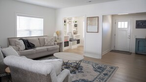 Un salon comprenant un canapé, un fauteuil, une table basse et un tapis. Une porte donne accès à une autre pièce où se trouvent des étagères et un meuble.