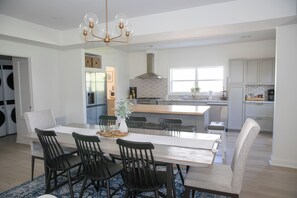 A modern kitchen with a central island, a dining area with a long table and chairs, and a large window providing natural light.