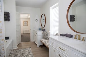 A bedroom with a bed, a dresser, a vanity with a mirror, and a door leading to another room.