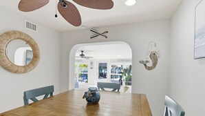 Een eetgedeelte met een houten tafel, stoelen, een geweven spiegel en een plafondventilator.