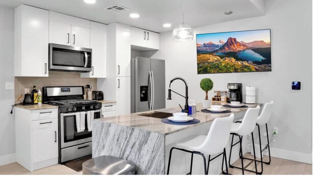 Una cocina moderna con armarios blancos, una isla central con taburetes de bar, un lavamanos grande en la isla y un cuadro de un paisaje montañoso.