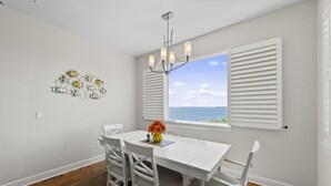 A dining area with a white table, chairs, a chandelier, and a view of the sea through a window with white shutters. - TOP FLORIDA's Villa Porpoise Point (Cape Coral)