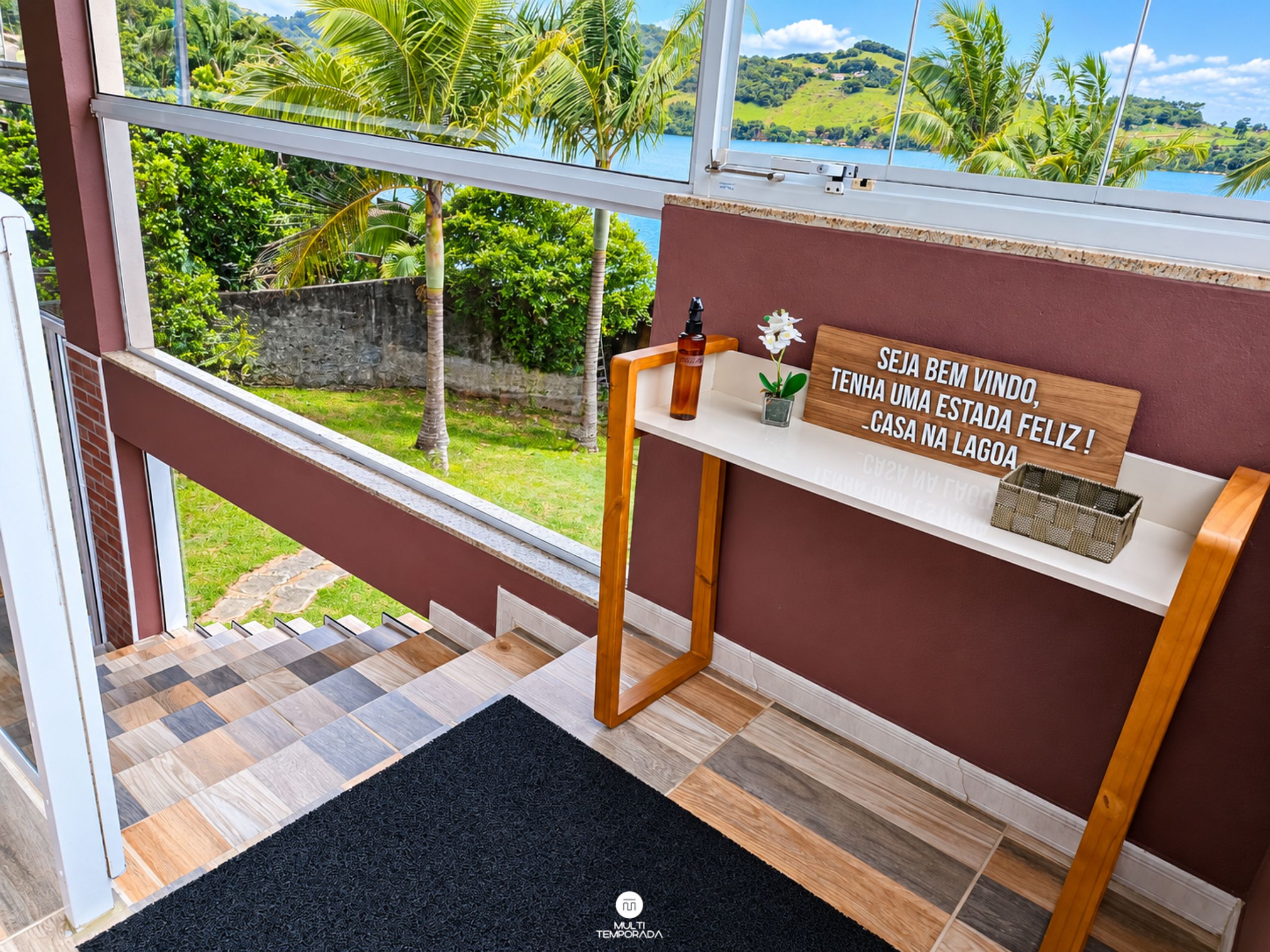 A room with a view of a lake, a wooden shelf with a bottle and a sign, and a patterned floor.