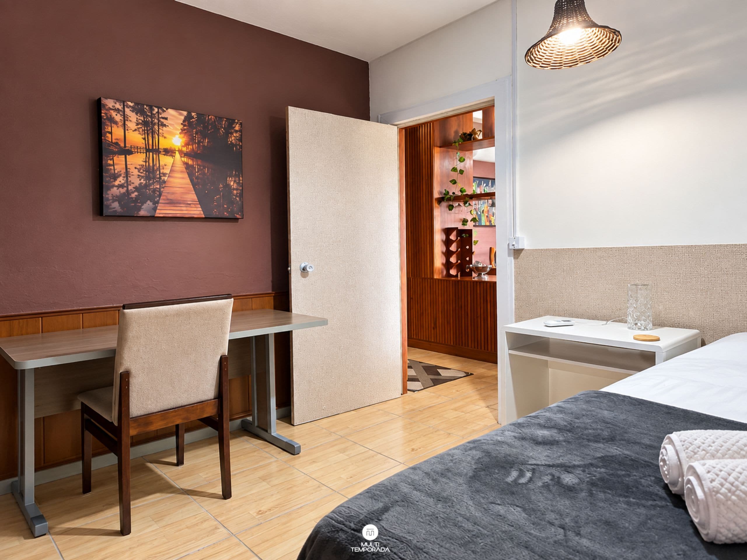 Hotellihuoneessa on sänky, työpöytä ja tuoli. Seinällä on taulu ja ovi johtaa toiseen huoneeseen.