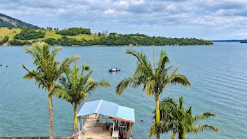 A small house with a roof and a dock extending into a calm lake, surrounded by palm trees and a hilly landscape.