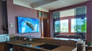 A kitchen with granite countertops, a microwave, a sink, a large flat-screen TV, and a view of a mountainous landscape through large windows. - Casa na Lagoa Imaruí-SC (Imaruí)