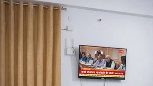 A room with a television mounted on the wall displaying a news broadcast, a brown leather sofa, a potted plant, and a curtain. - 2-bedroom apartment in perfect Patna with cool AC (Patna)