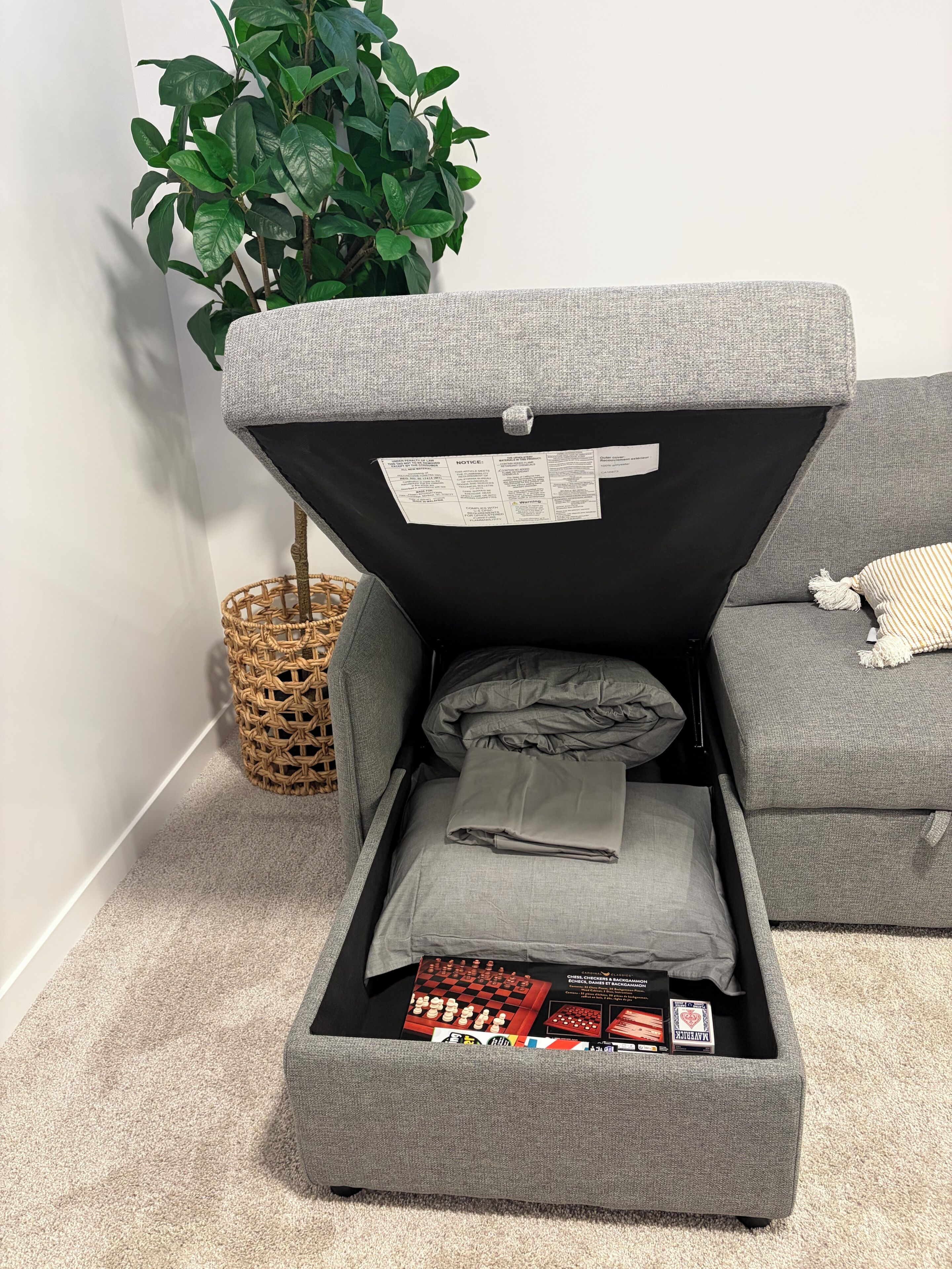 A grey sofa with a storage compartment underneath, a woven basket, and a green plant.