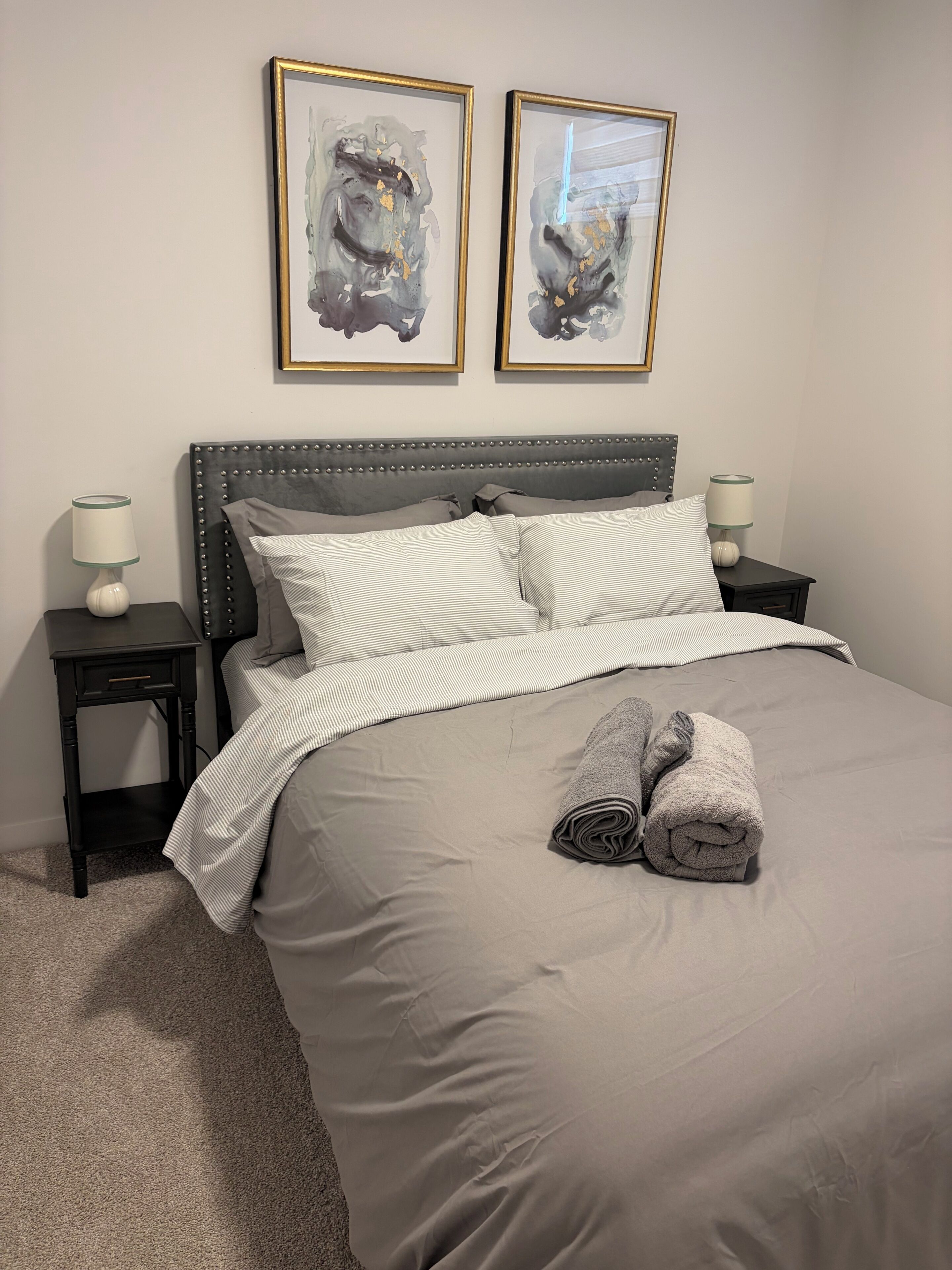 A neatly made bed with striped bedding, two bedside lamps, and two framed abstract paintings on the wall.