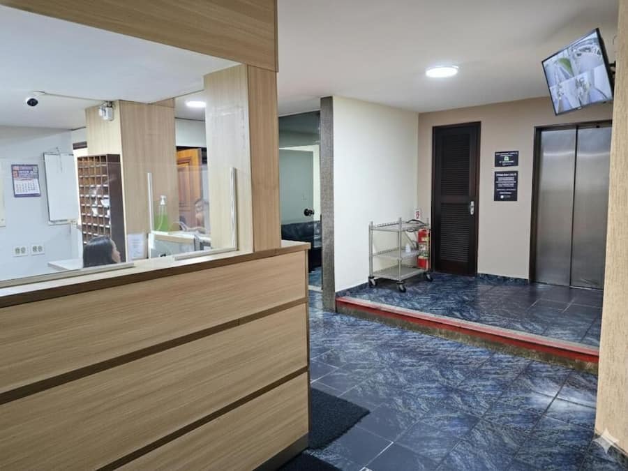 Uma área de recepção com balcão de madeira, piso ladrilhado, televisão na parede e uma porta com placa.