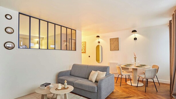 Un salon moderne avec un canapé gris, une table de salle à manger ronde entourée de chaises, une petite table basse et un grand miroir accroché au mur.