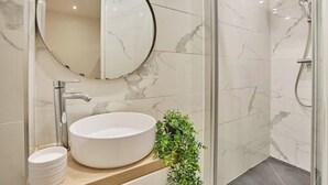 Une salle de bain avec un miroir rond, un lavabo blanc, une douche et une petite plante verte.