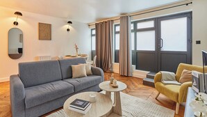 Un salon moderne avec un canapé gris, un fauteuil jaune, une table basse et une grande fenêtre avec des rideaux.