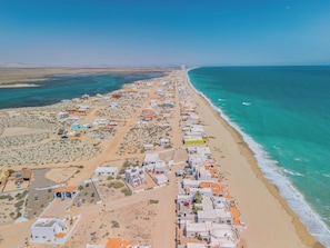 An aerial view of a coastal area with buildings aligned along the beachfront, a clear sky, and a calm sea. - Casa La Paz|Beachfront in Playa La Jolla|10 Guests (Playa Encanto)